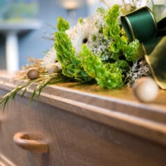 A close-up of a wooden coffin adorned with a floral arrangement featuring white flowers and green ribbon, with soft lighting in the background—evoking the gentle care of a compassionate Funeral Director.