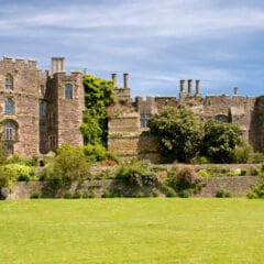 A large stone castle with multiple chimneys, arched windows, and leafy ivy growing on the walls stands behind a lush green lawn and flowering shrubs under a bright blue sky—the perfect peaceful setting for woodland burials and reflecting on funeral costs.