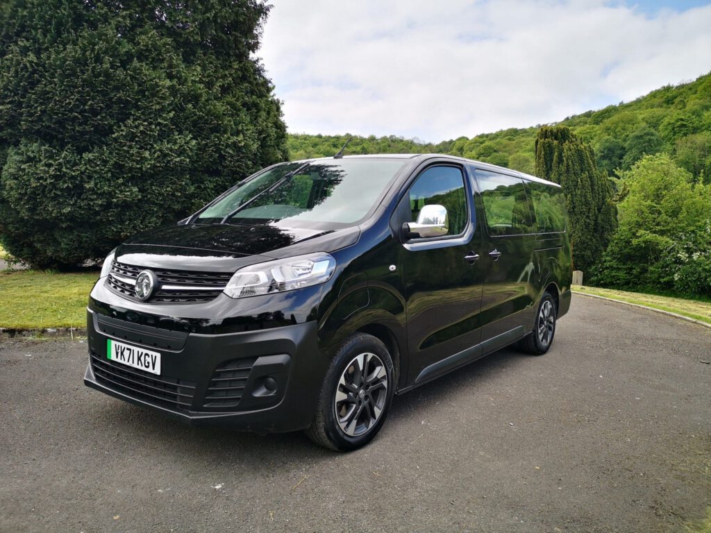 A black Vauxhall van is parked on a paved surface surrounded by greenery and trees, with hills and a partly cloudy sky in the background.