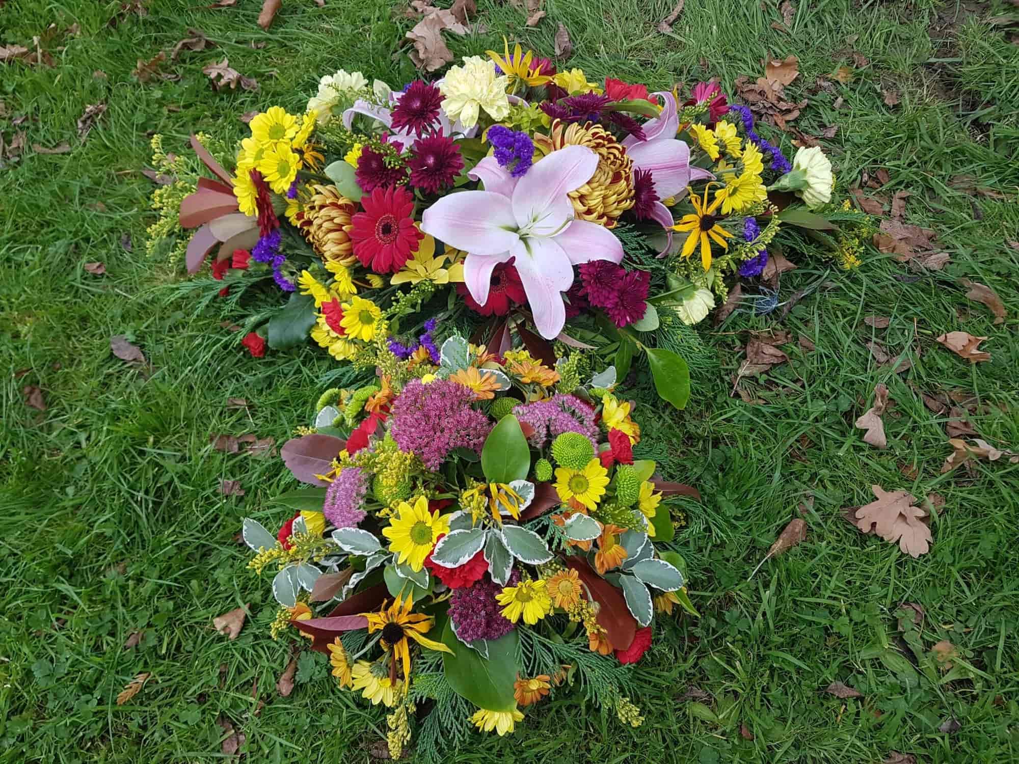 Two colorful floral arrangements lie on green grass. The bouquets feature lilies, chrysanthemums, daisies, and various other flowers in pink, yellow, red, and purple hues, surrounded by green foliage and scattered fallen leaves nearby.