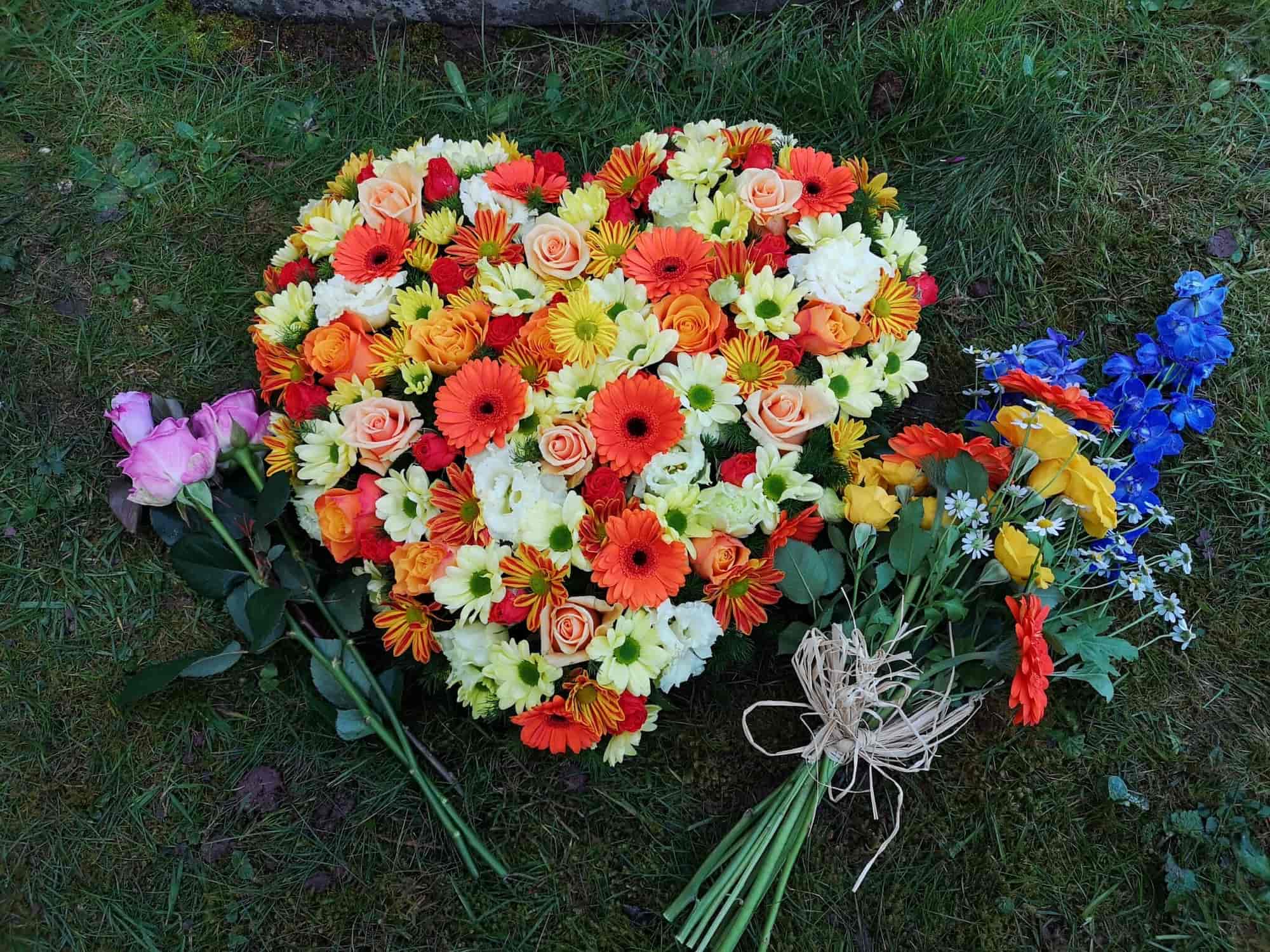 A heart-shaped floral arrangement of orange, yellow, and white flowers lies on grass, with additional bouquets of pink, blue, and orange flowers placed next to it.