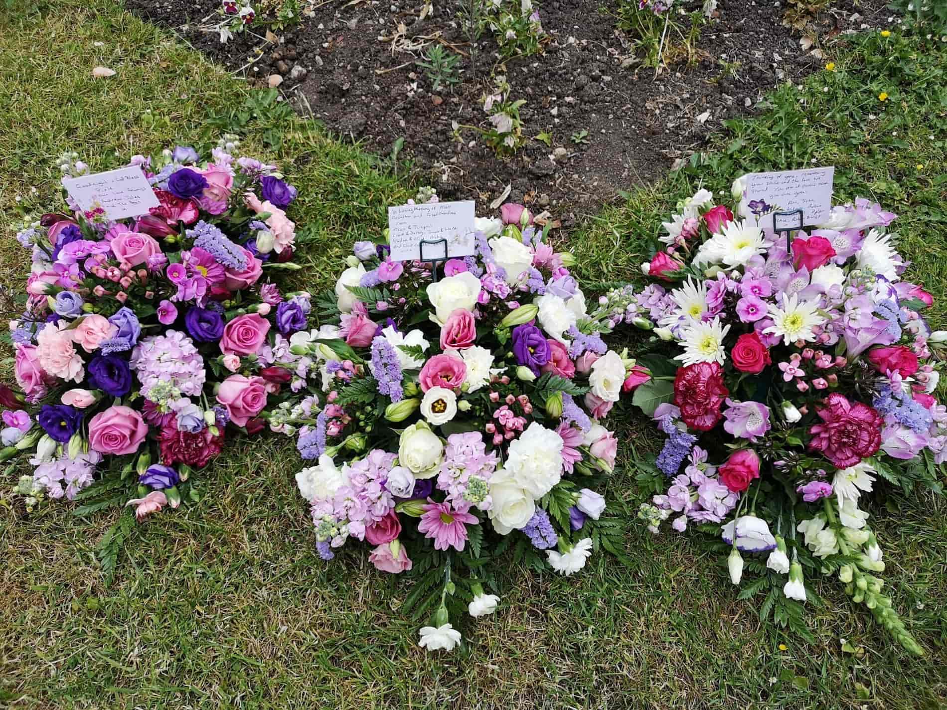 Three floral arrangements with pink, white, and purple flowers lie on grass in front of a garden bed. Each arrangement has a card attached, likely messages for a memorial or funeral.