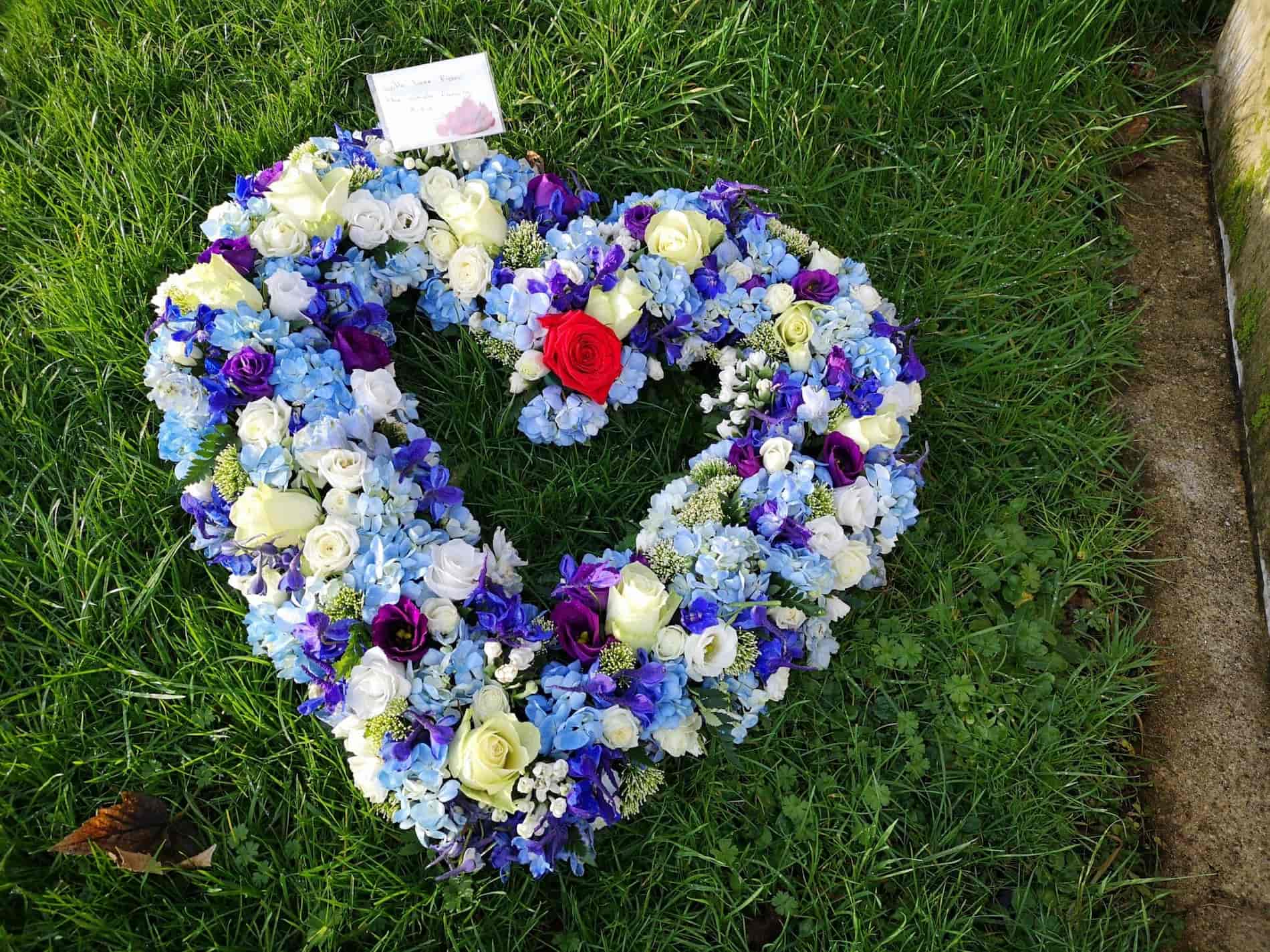 A heart-shaped floral wreath with white, blue, purple, and a single red rose, placed on green grass next to a card and a stone surface.