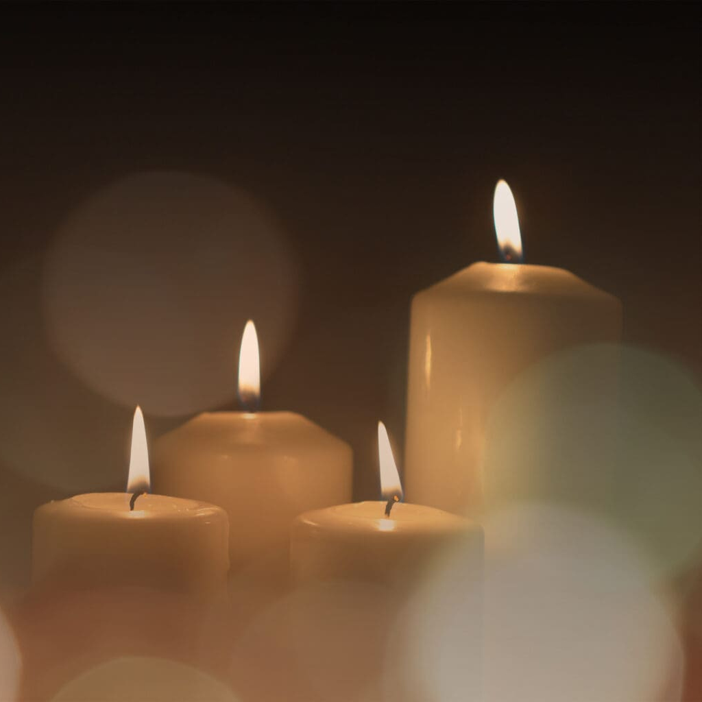 Four lit candles of varying heights are grouped together against a dark background, with soft, blurred light circles in the foreground, creating a warm and peaceful atmosphere.