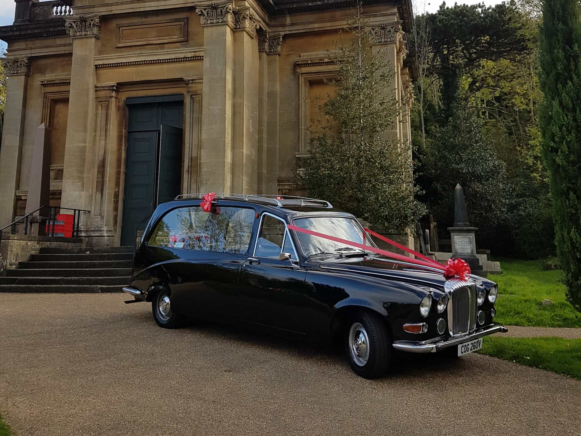 A black vintage hearse decorated with pink ribbons and bows is parked outside a stone building with columns, surrounded by greenery.