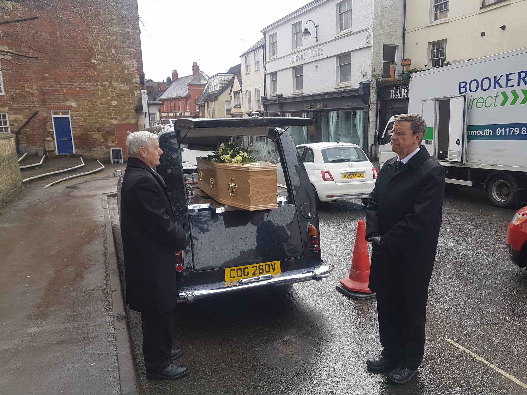 Two men in black suits stand near a vintage hearse with a coffin inside, parked on a street. A traffic cone and a delivery truck are visible in the background.