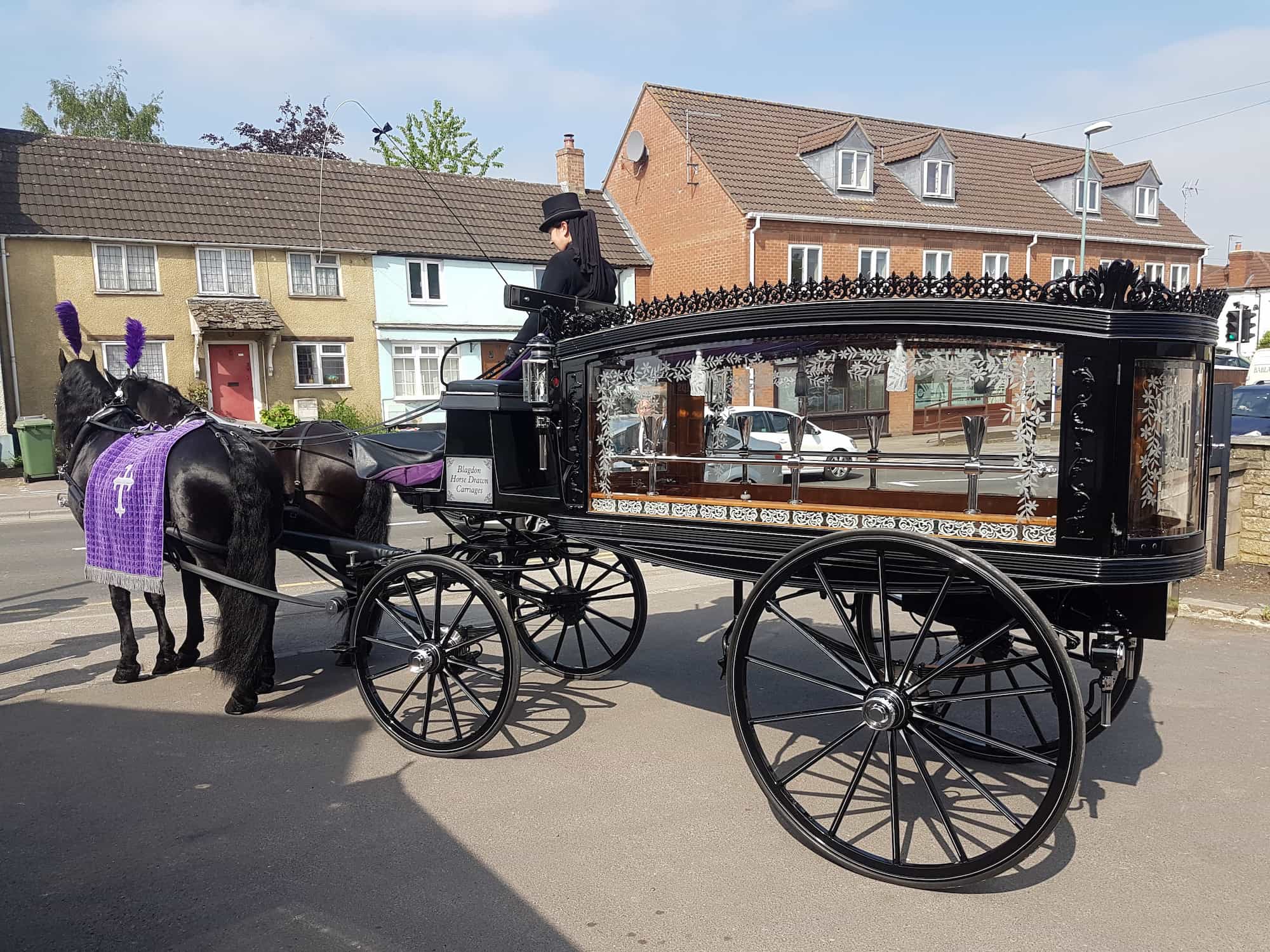 A black horse-drawn funeral hearse carriage with glass sides stands on a street; the horse is draped in a purple cloth, and the driver wears traditional formal attire.