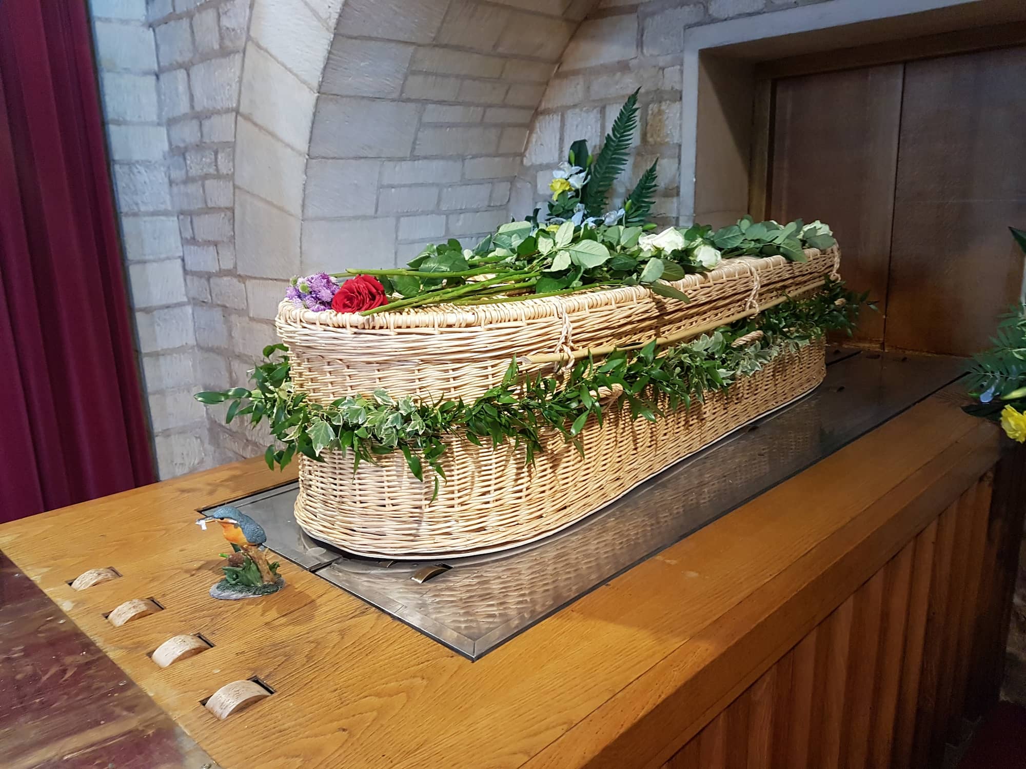 A wicker coffin adorned with green foliage and flowers rests on a wooden platform inside a stone-walled room, near a metal plaque decorated with a bird figure.