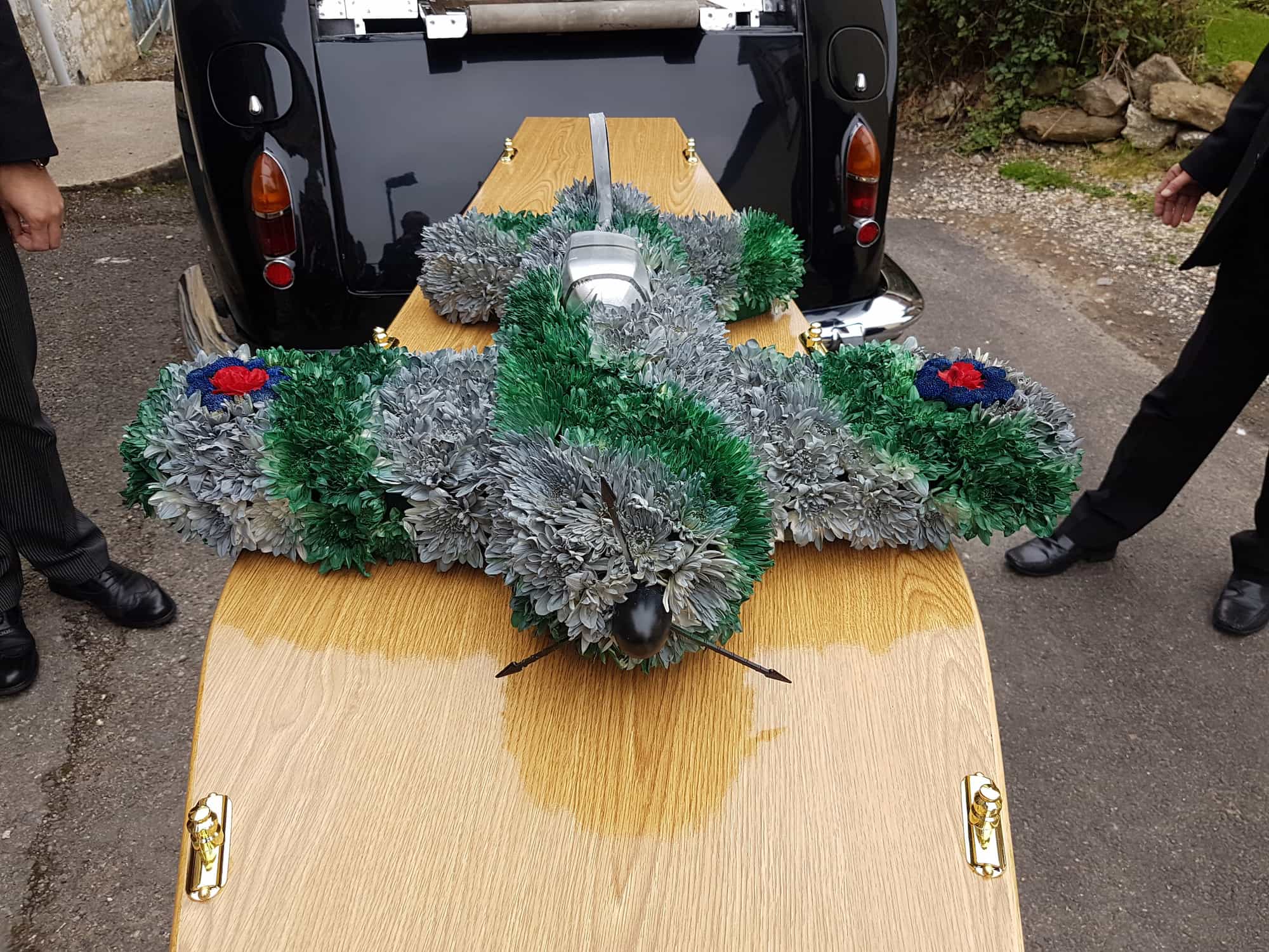 A floral arrangement shaped like an airplane sits on top of a wooden coffin, with two people standing nearby and a black vehicle in the background.