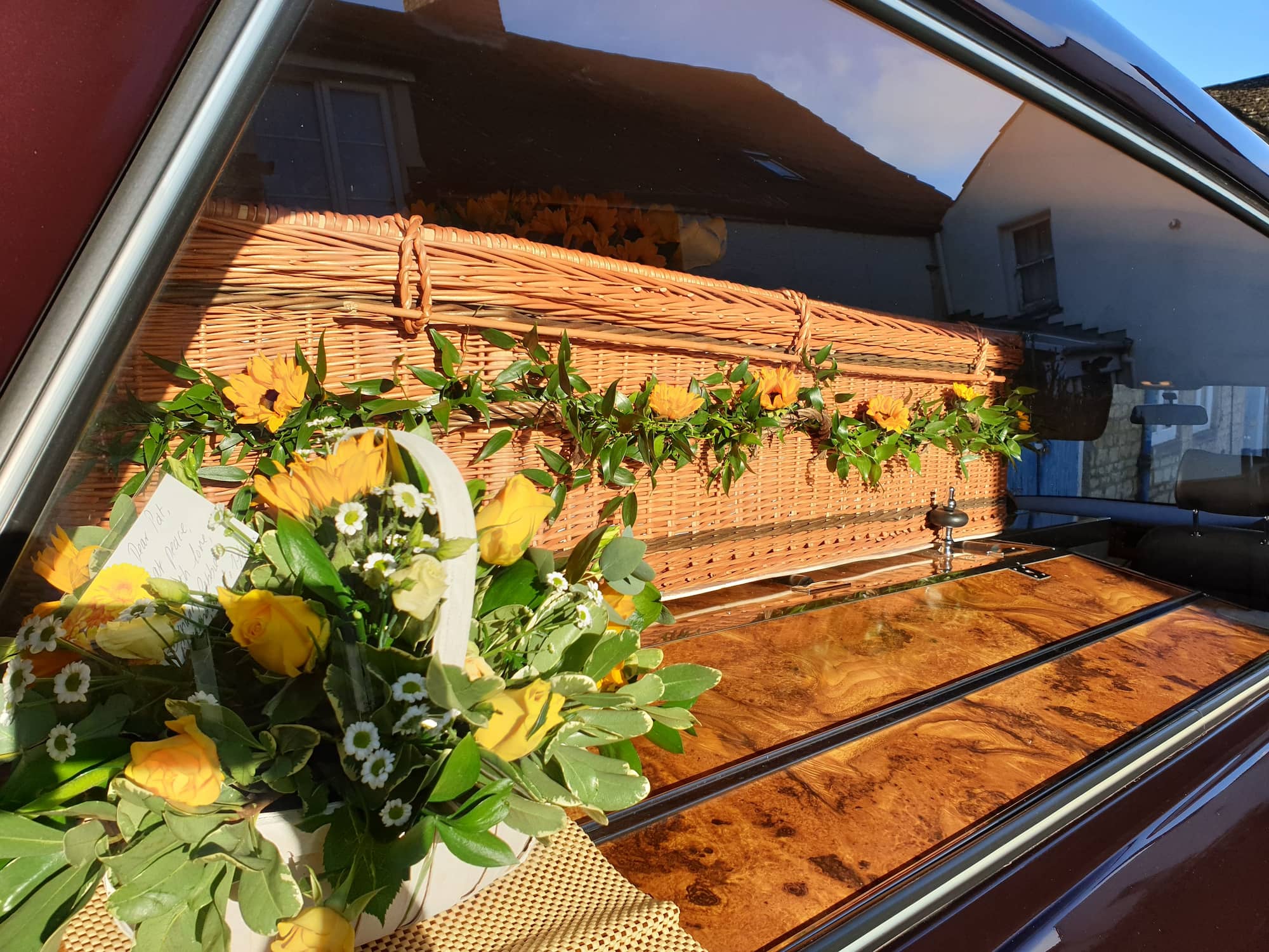 A wicker coffin adorned with yellow flowers is seen inside a vehicle, with a basket of yellow flowers and greenery placed beside it. Sunlight reflects on the window, showing buildings outside.