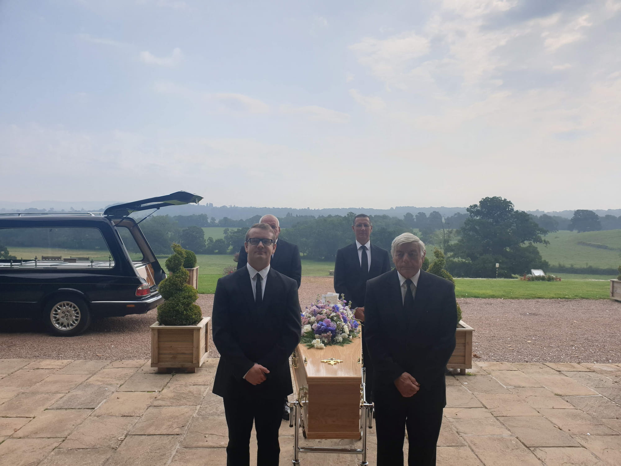 Four men in black suits stand by a wooden coffin adorned with flowers outside, with a hearse and green fields visible in the background under a cloudy sky.