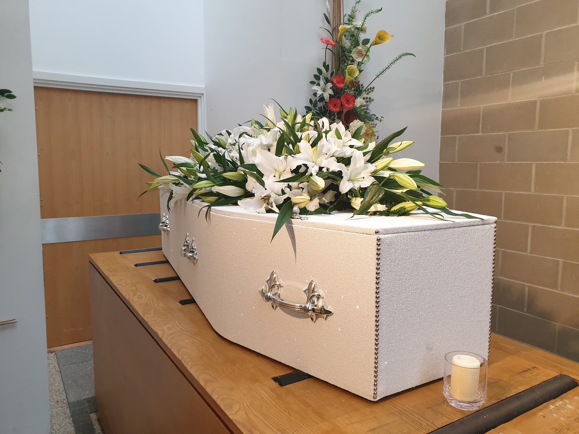 A white coffin adorned with white lilies rests on a wooden stand. A small candle is placed beside it, and a floral arrangement is displayed in the background.