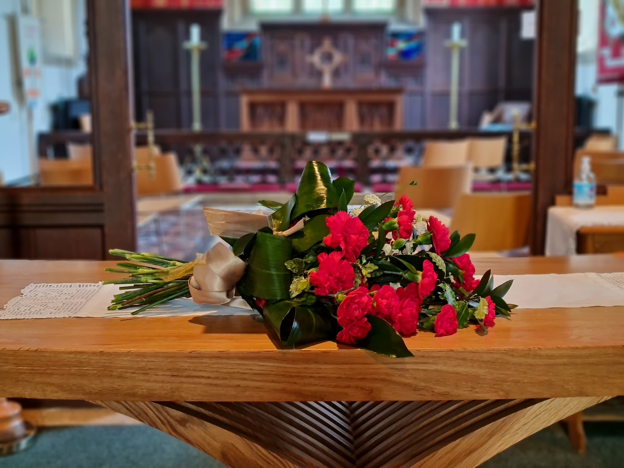 A bouquet of red flowers rests on a wooden table inside a church, with pews, an altar, and a cross visible in the blurred background.