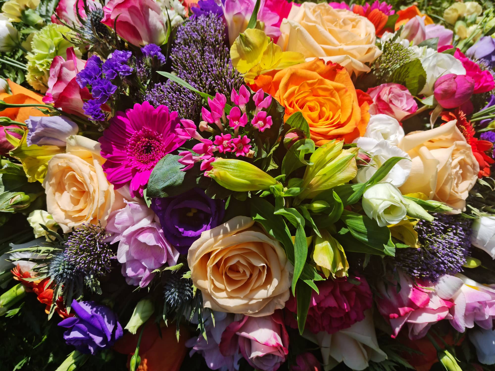A colorful bouquet of mixed flowers, including roses, gerbera daisies, and various other blossoms in shades of pink, orange, yellow, purple, and white, arranged closely together.