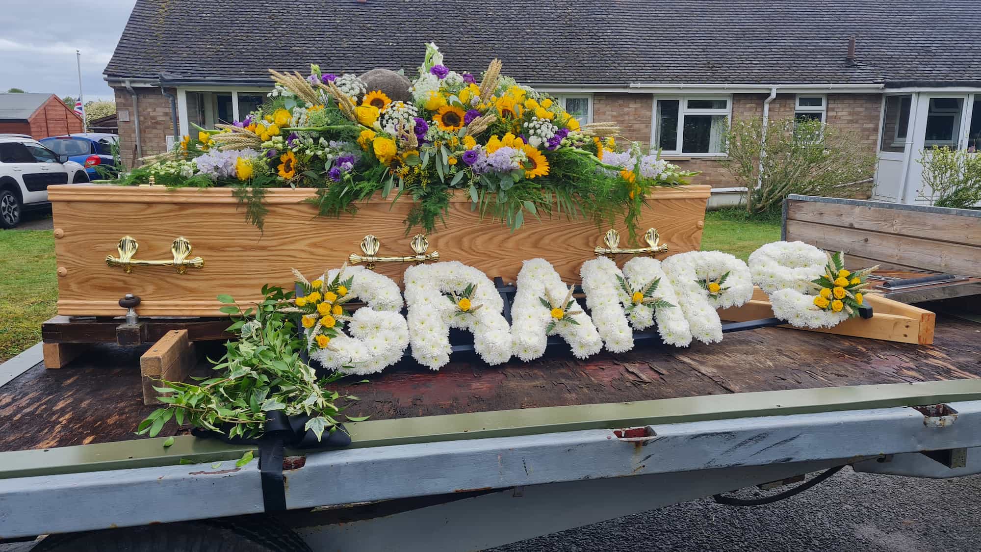 A wooden coffin adorned with a large, colorful flower arrangement and white floral letters spelling "GRAMP" rests on a flatbed trailer outside a brick building.