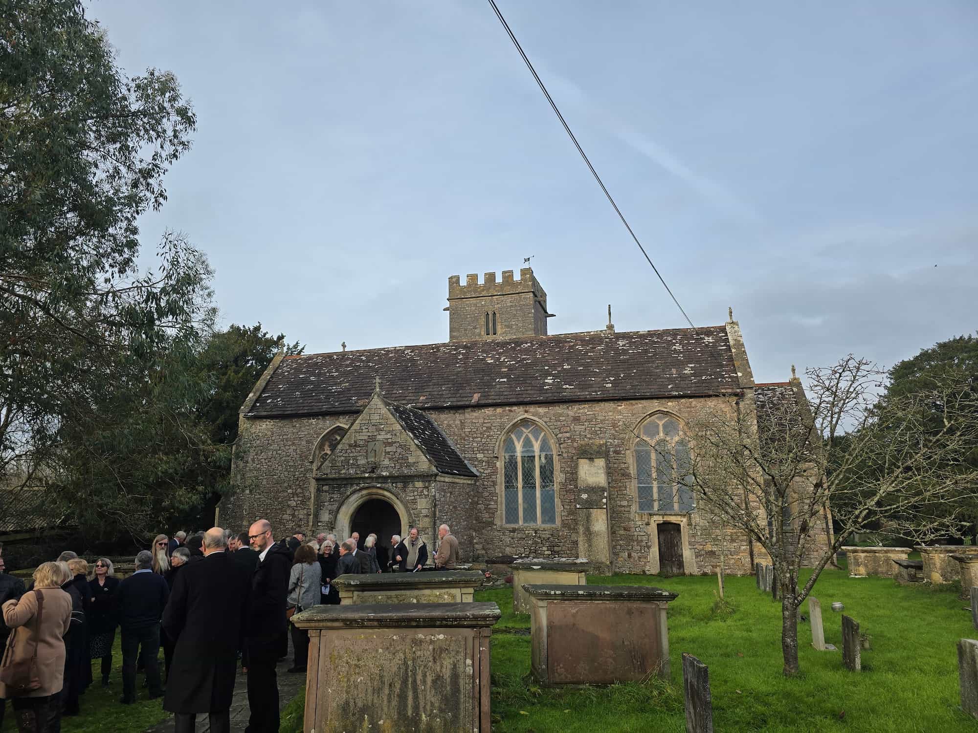 A group of people gather outside an old stone church with a tower and arched doorway, surrounded by gravestones and green grass under a cloudy sky.