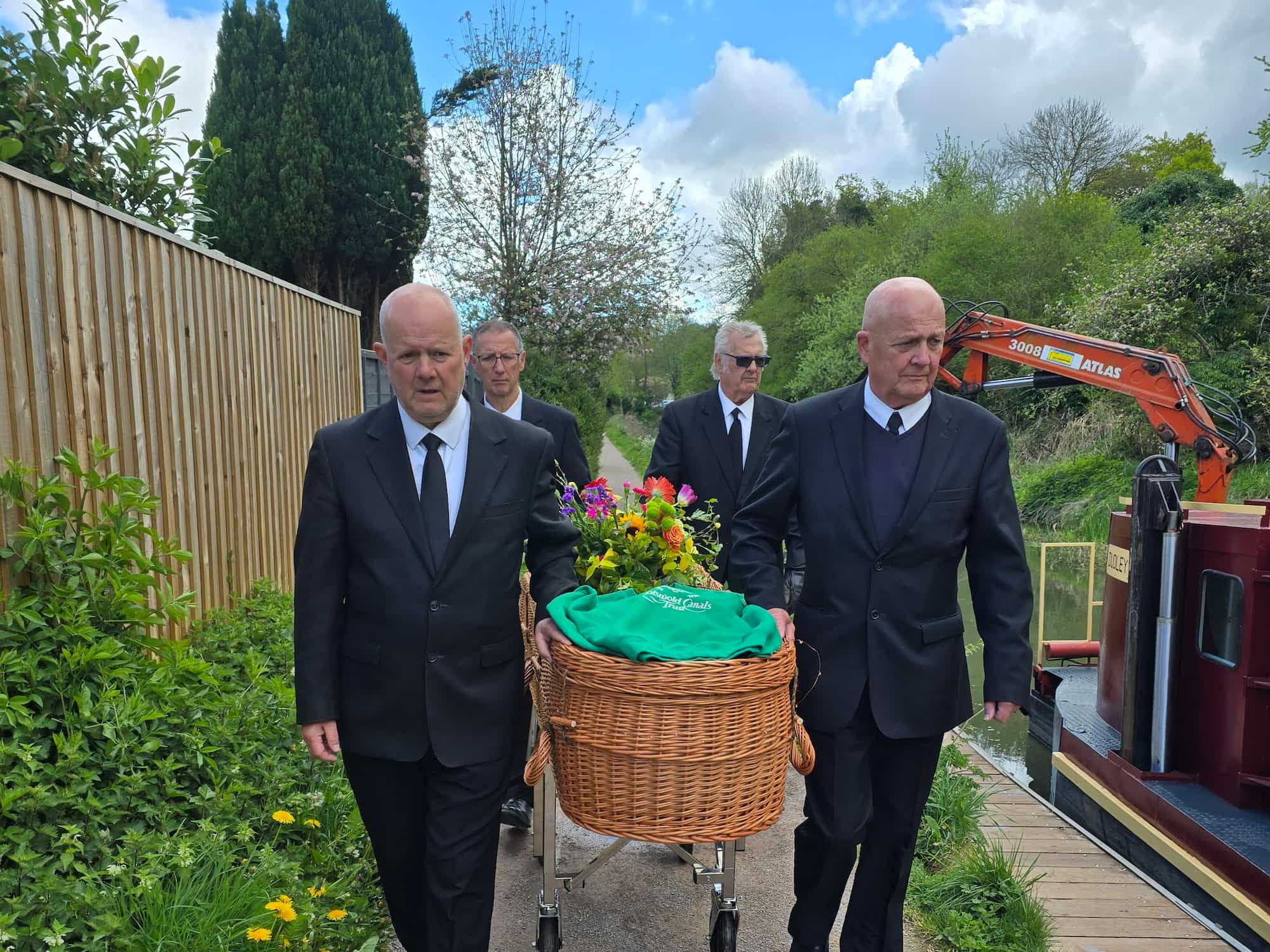Four men in black suits carry a wicker coffin adorned with colorful flowers along a garden path beside a wooden fence, with greenery and construction equipment in the background.
