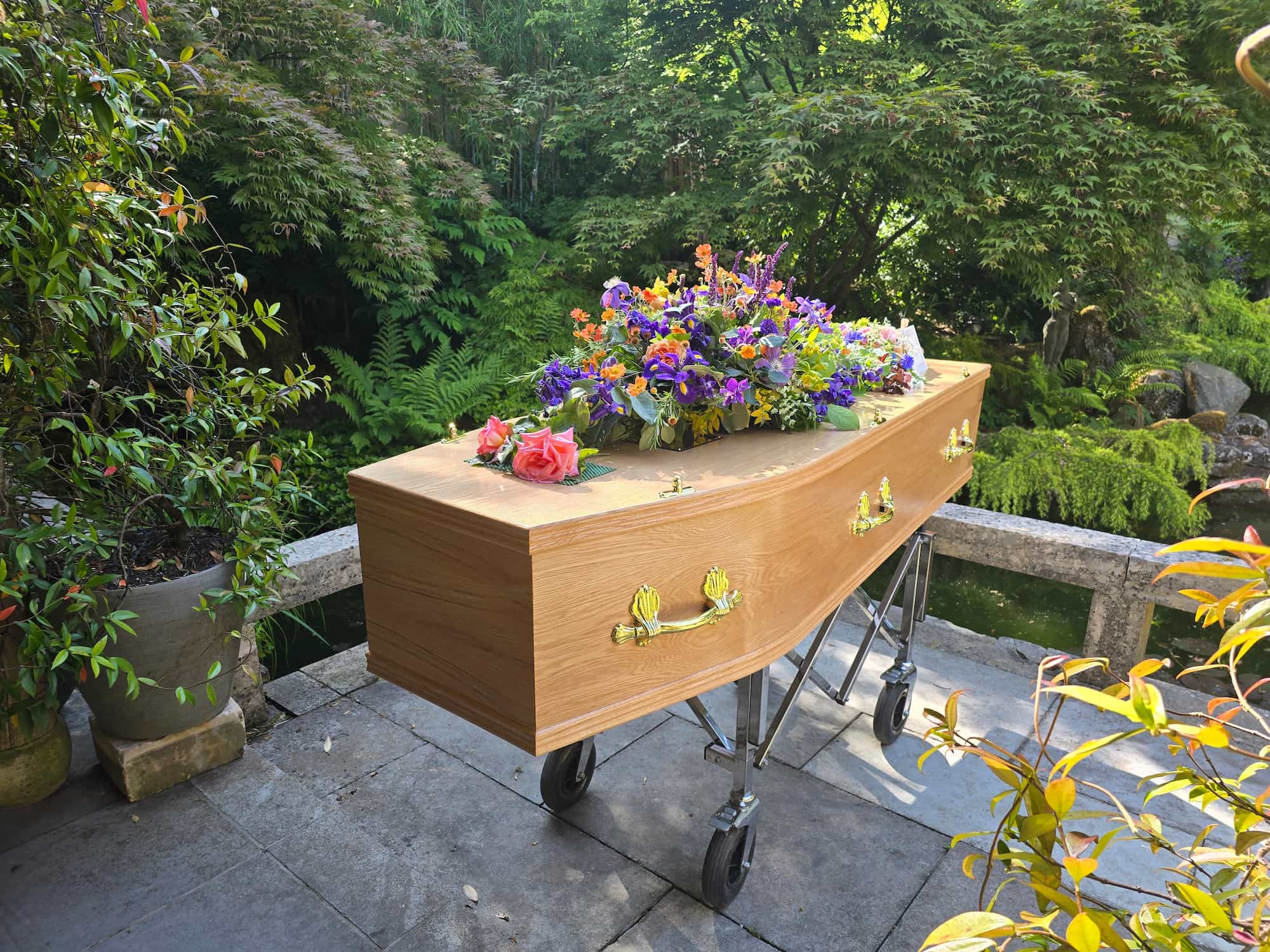 A wooden coffin with gold handles and colorful flowers on top stands on a trolley outdoors, surrounded by green plants and trees.