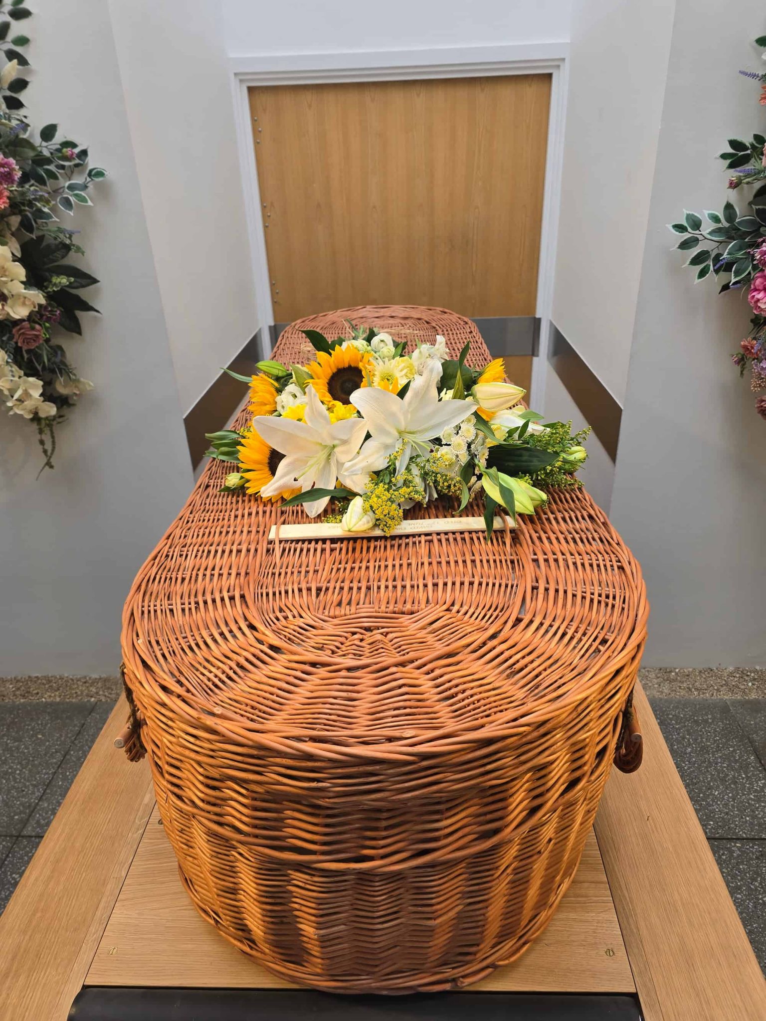 A wicker coffin topped with a bouquet of white lilies and yellow sunflowers is placed in a bright room with floral arrangements on the walls.