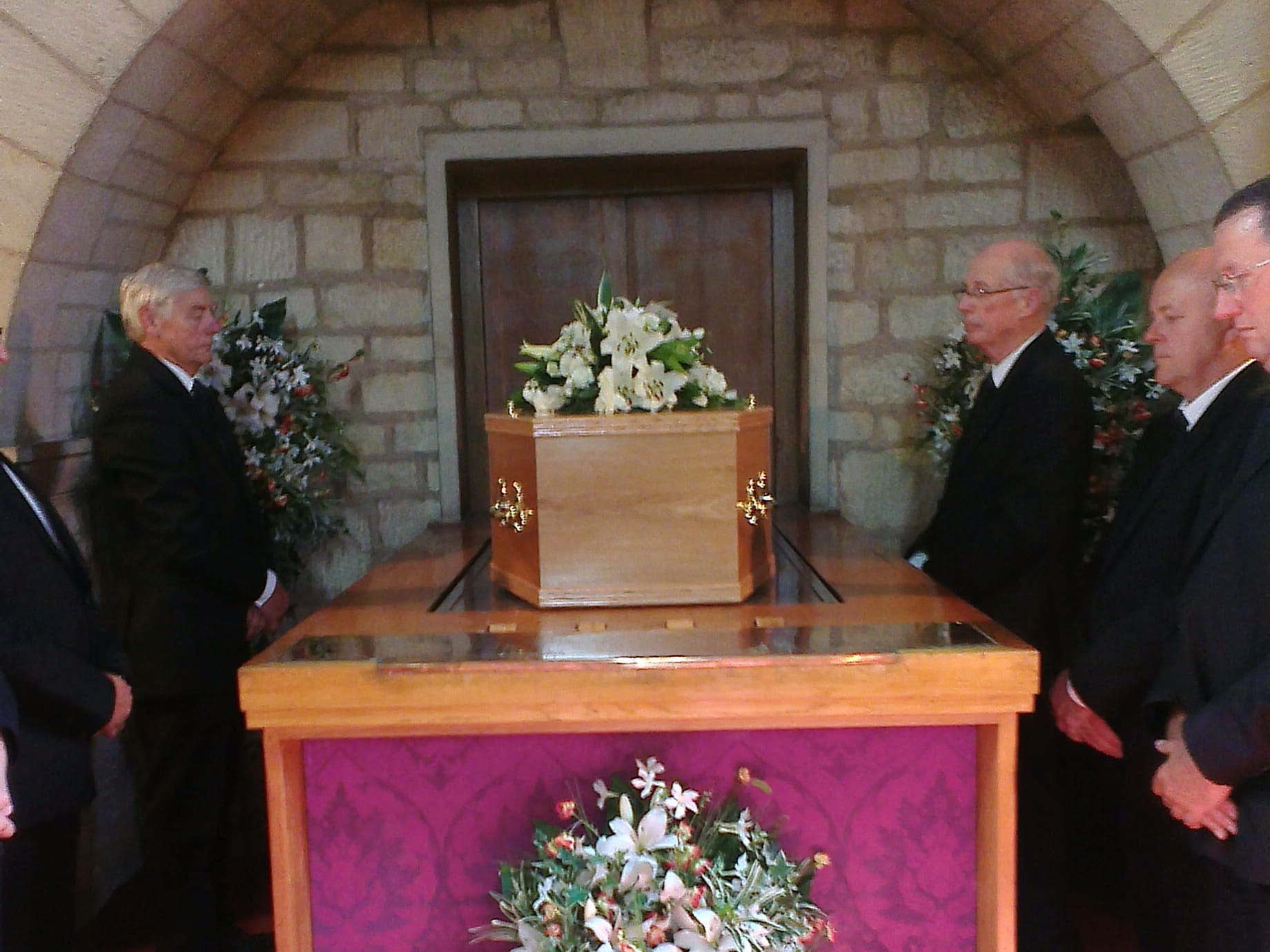 Four men in black suits stand around a wooden casket adorned with white flowers inside a stone-walled room, with floral arrangements nearby.