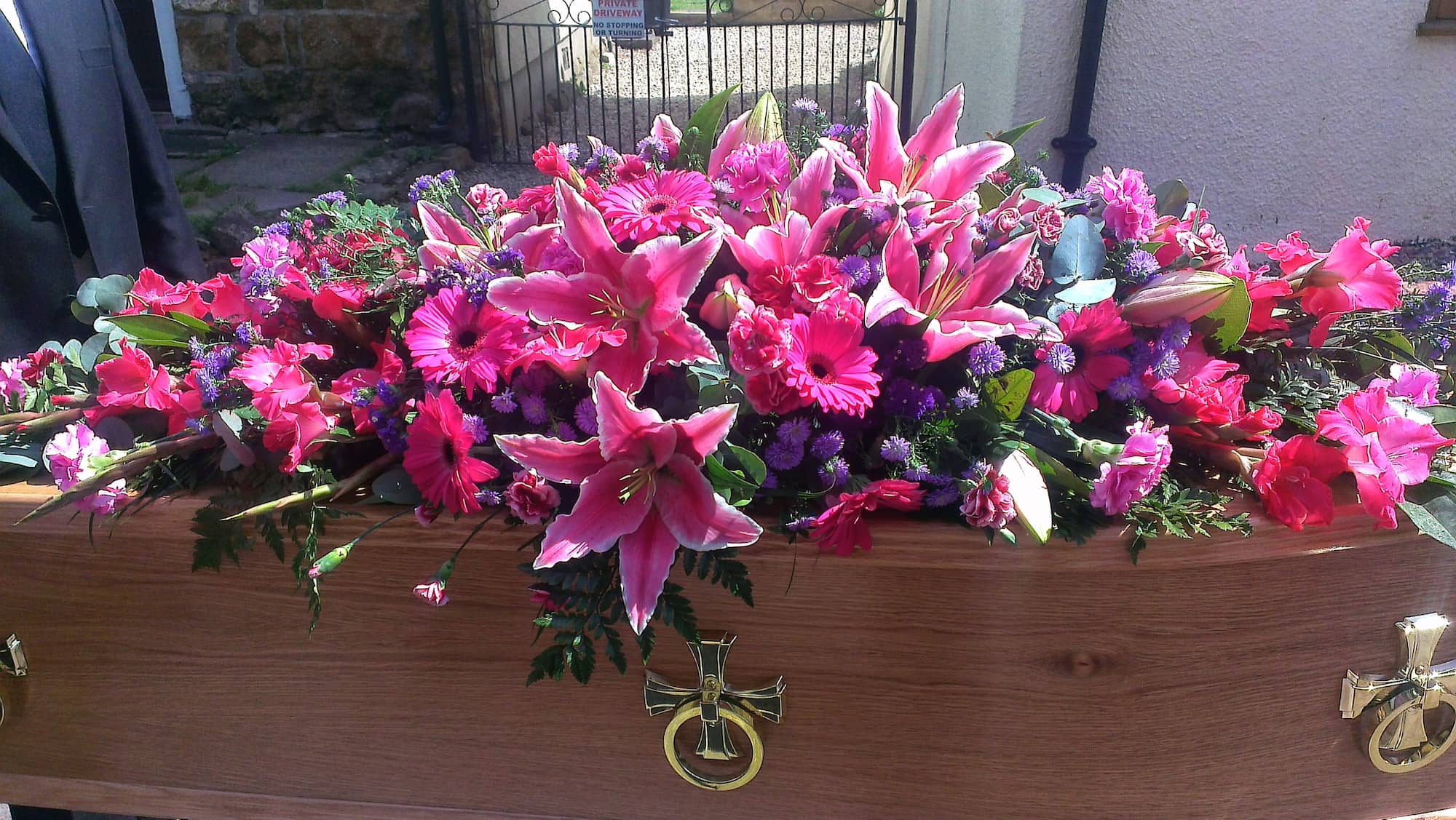 A wooden coffin adorned with a large, colorful arrangement of pink lilies, purple and pink flowers, and greenery, outdoors near a gate.