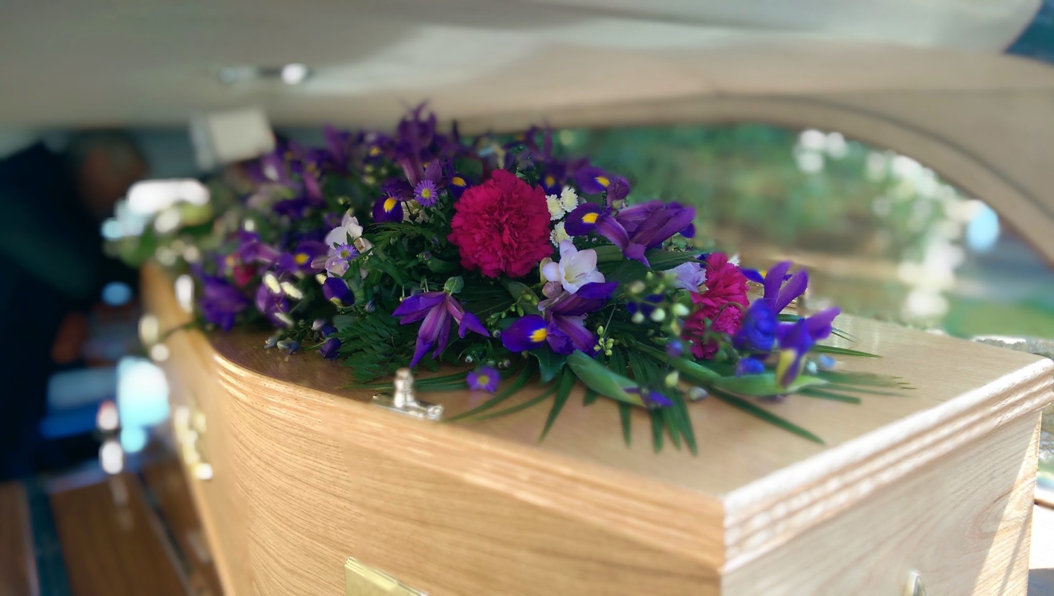 A wooden coffin with a large floral arrangement of purple and pink flowers on top, inside a vehicle, likely a hearse.