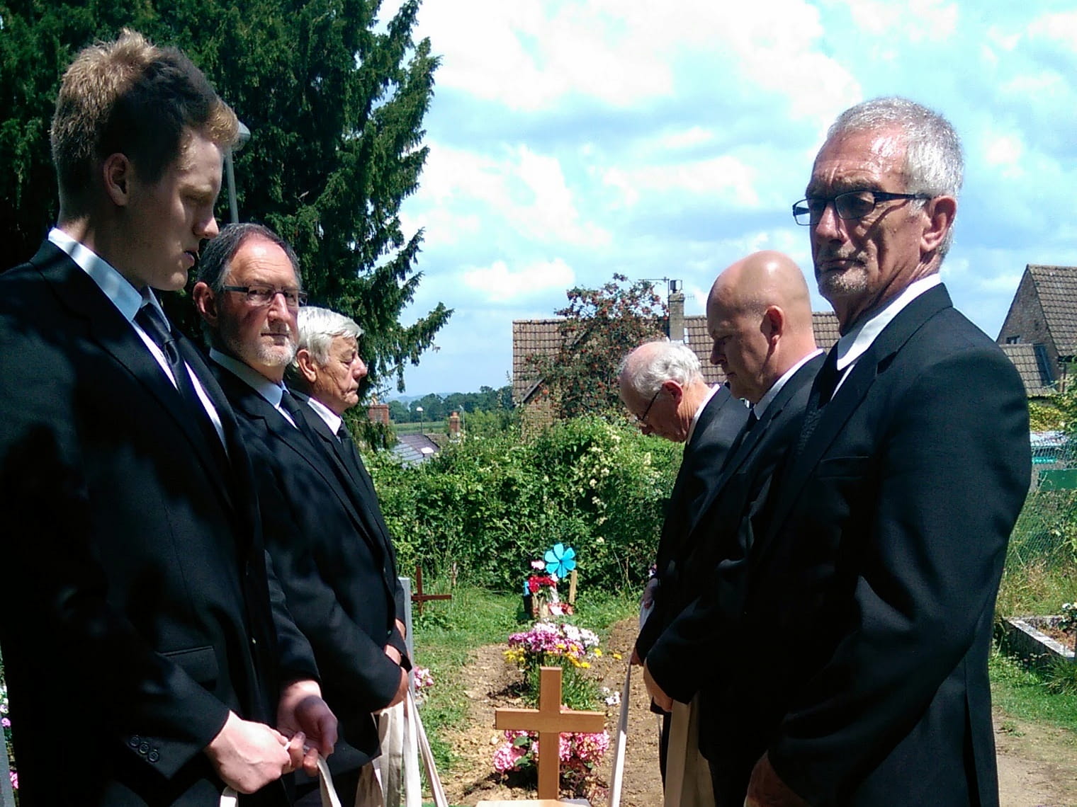 Six men in black suits stand in two lines facing each other at a gravesite, heads bowed and solemn, with flowers and a wooden cross marking the grave. Trees and houses are visible in the background.