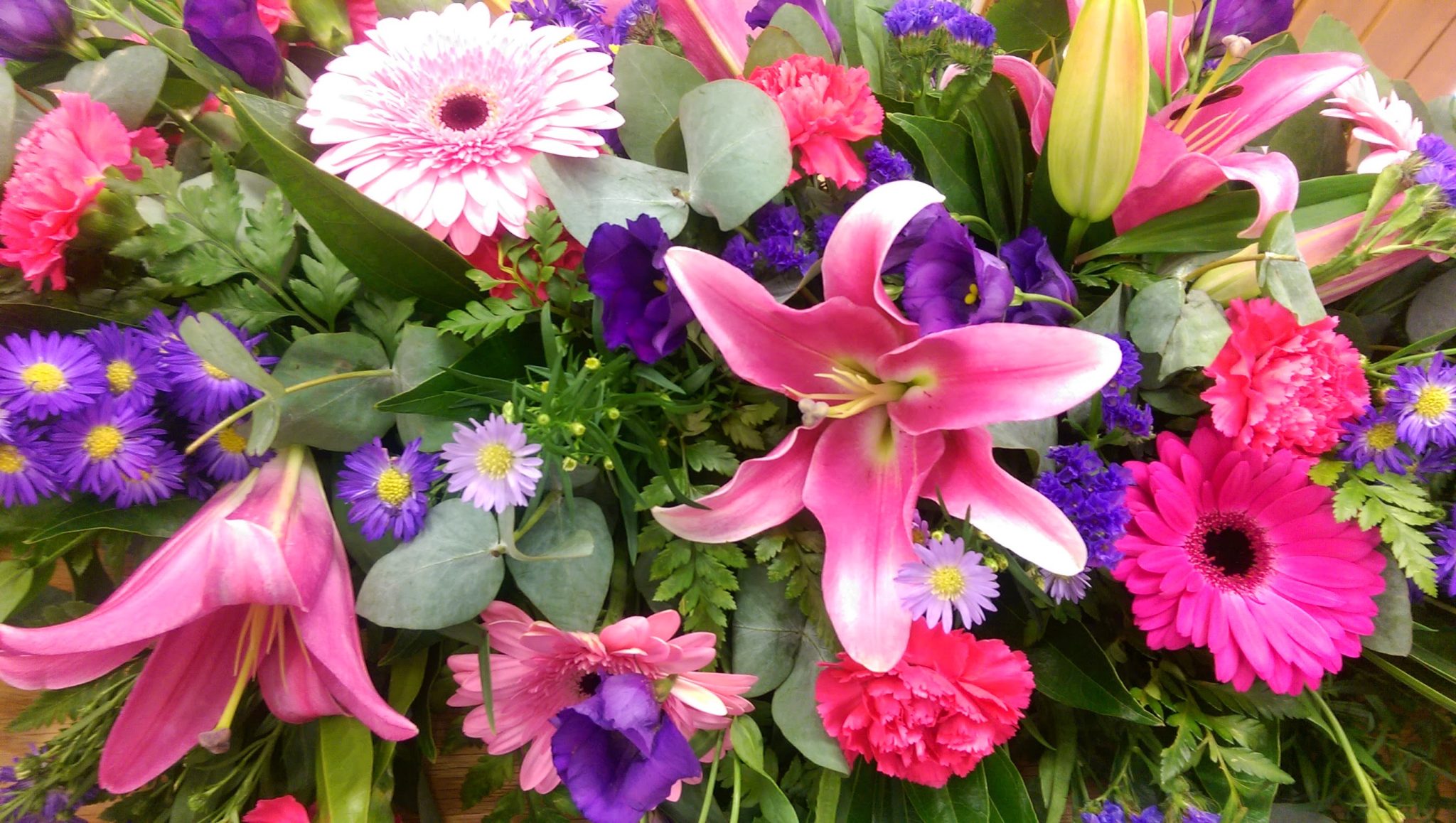 A vibrant bouquet featuring pink lilies, gerbera daisies, carnations, purple asters, and greenery. The flowers are arranged closely together, creating a lush and colorful display.