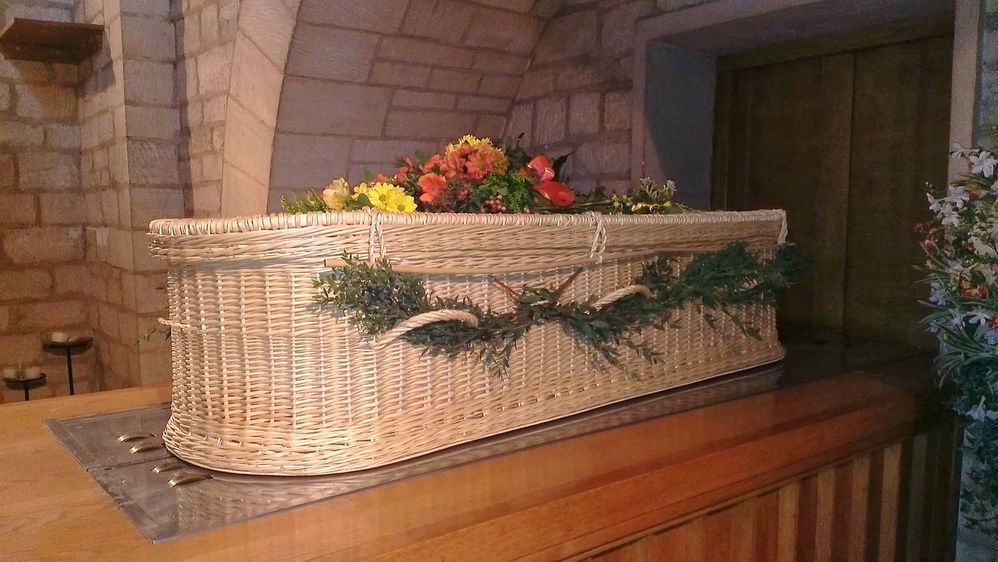 A wicker coffin decorated with greenery and topped with colorful flowers rests inside a stone-walled room, possibly a church or chapel.