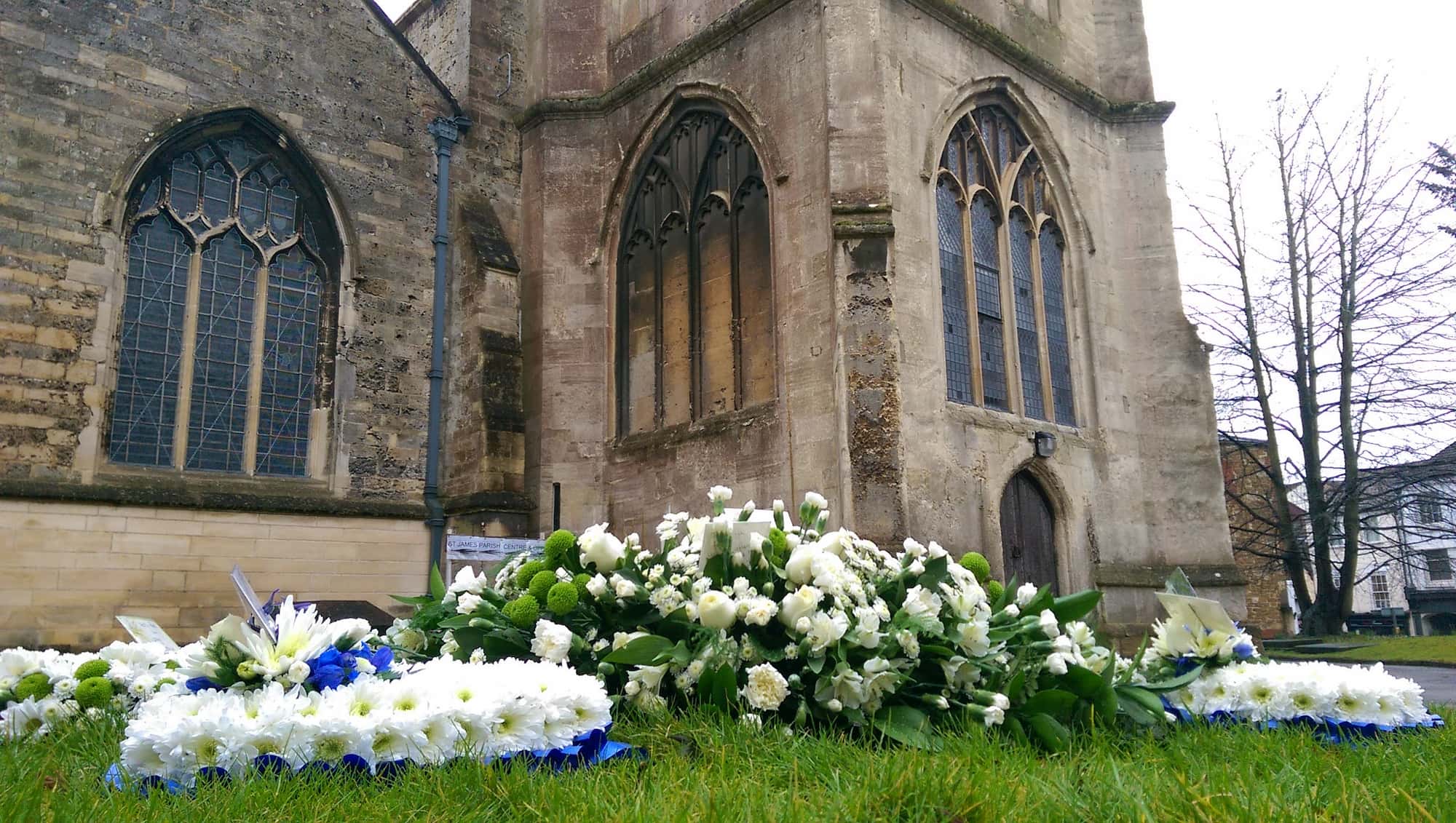 White and blue floral wreaths lie on grass outside a stone church with tall arched windows. The church has weathered walls and leafless trees are visible in the background.
