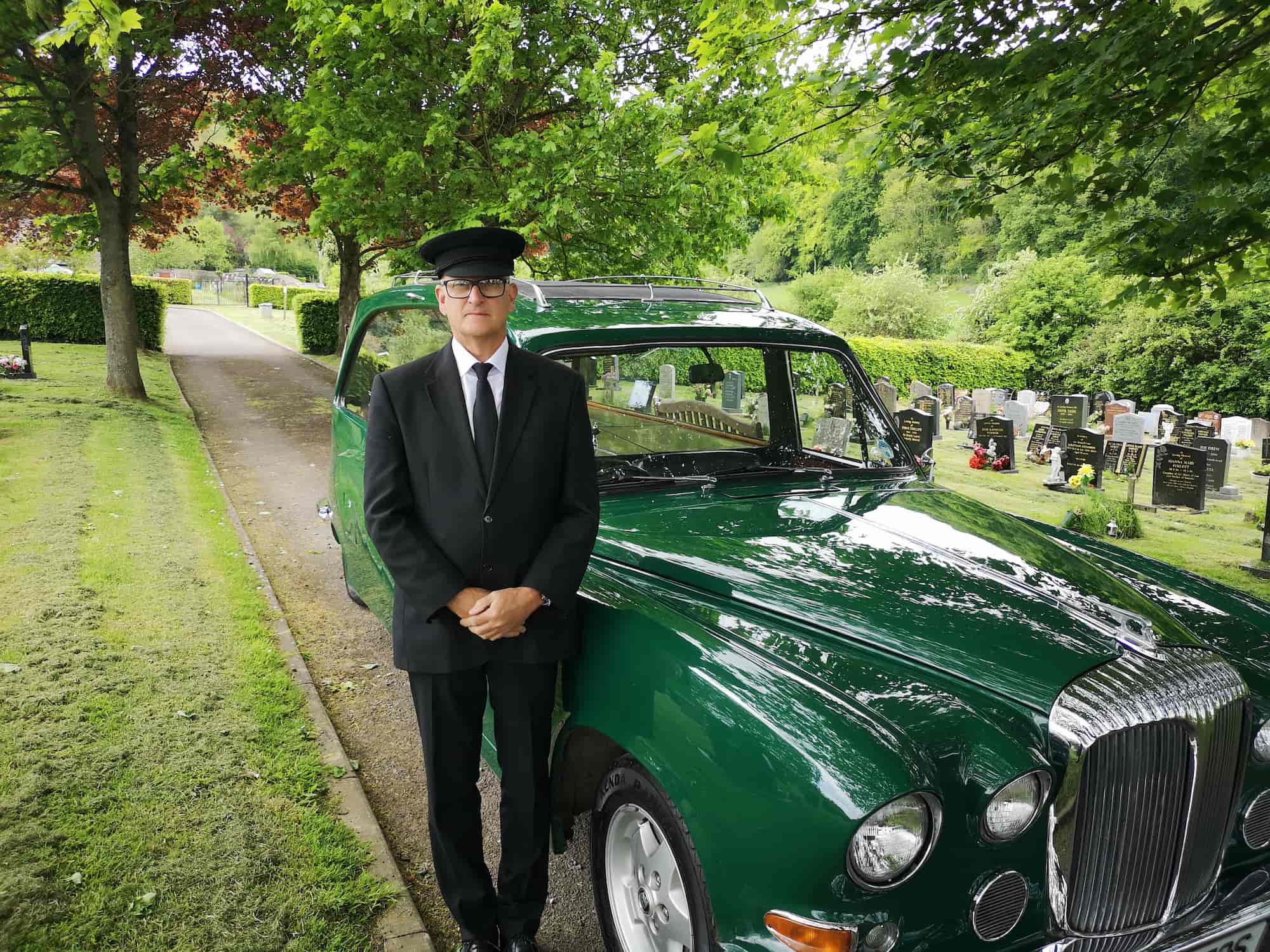 A man in a black suit and hat stands in front of a vintage green car in a cemetery, with gravestones and trees in the background.
