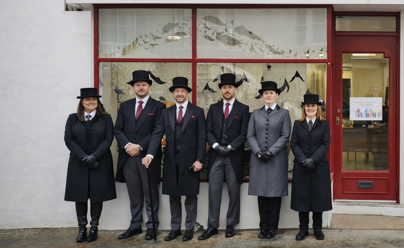 Six people wearing formal attire, including black coats, vests, ties, and top hats, stand in a row outside a building with a red-framed window and door. The group smiles for the photo on a wet sidewalk.