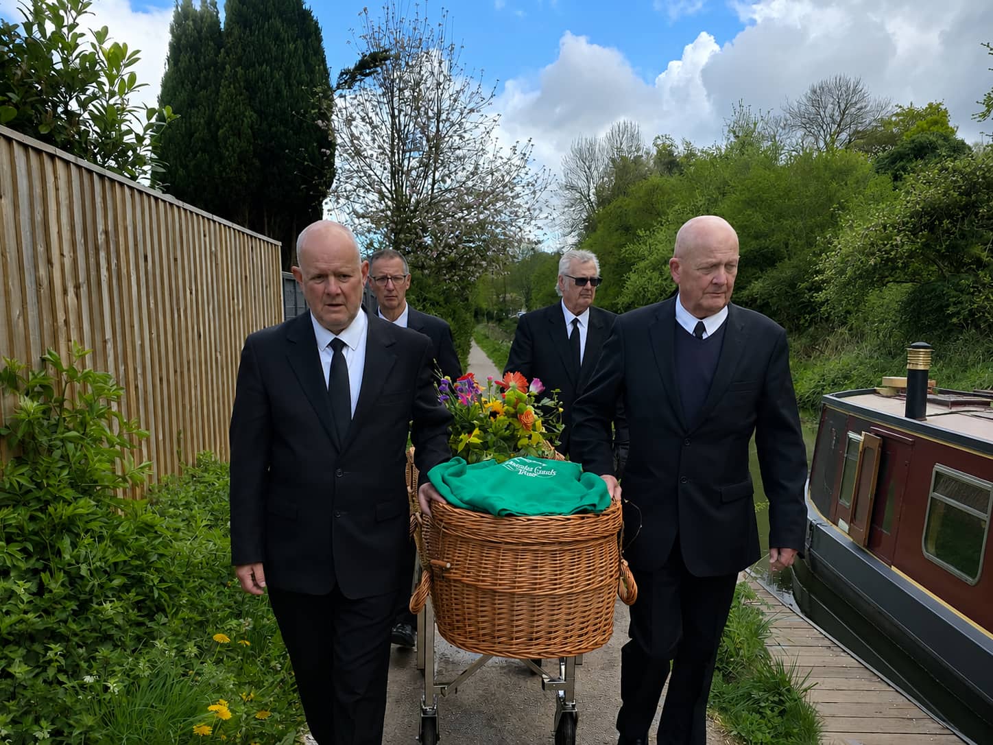 Four men in black suits carry a wicker casket with flowers along a path beside a canal boat, surrounded by greenery and a wooden fence under a partly cloudy sky.